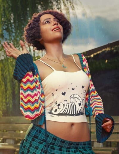 Vibrant young woman outdoors in a colorful zip-up hoodie with rainbow stripes, wearing a playful black-and-white cat graphic crop top, enjoying a rainy day under a cloudy sky, showcasing stylish fashion and expressive personality.