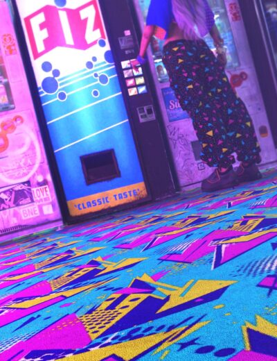 Vibrant neon-lit alley scene featuring a woman in colorful patterned pants standing next to a brightly illuminated vending machine with graffiti art. The scene captures a retro-futuristic, cyberpunk aesthetic with vivid colors and bold patterns.