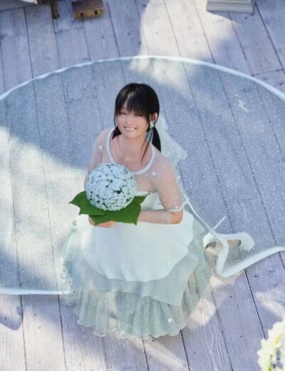 Beautiful young woman in a white dress holding a bouquet of white flowers, sitting on a white curved bench outdoors on a sunny day, celebrating a wedding or special event.
