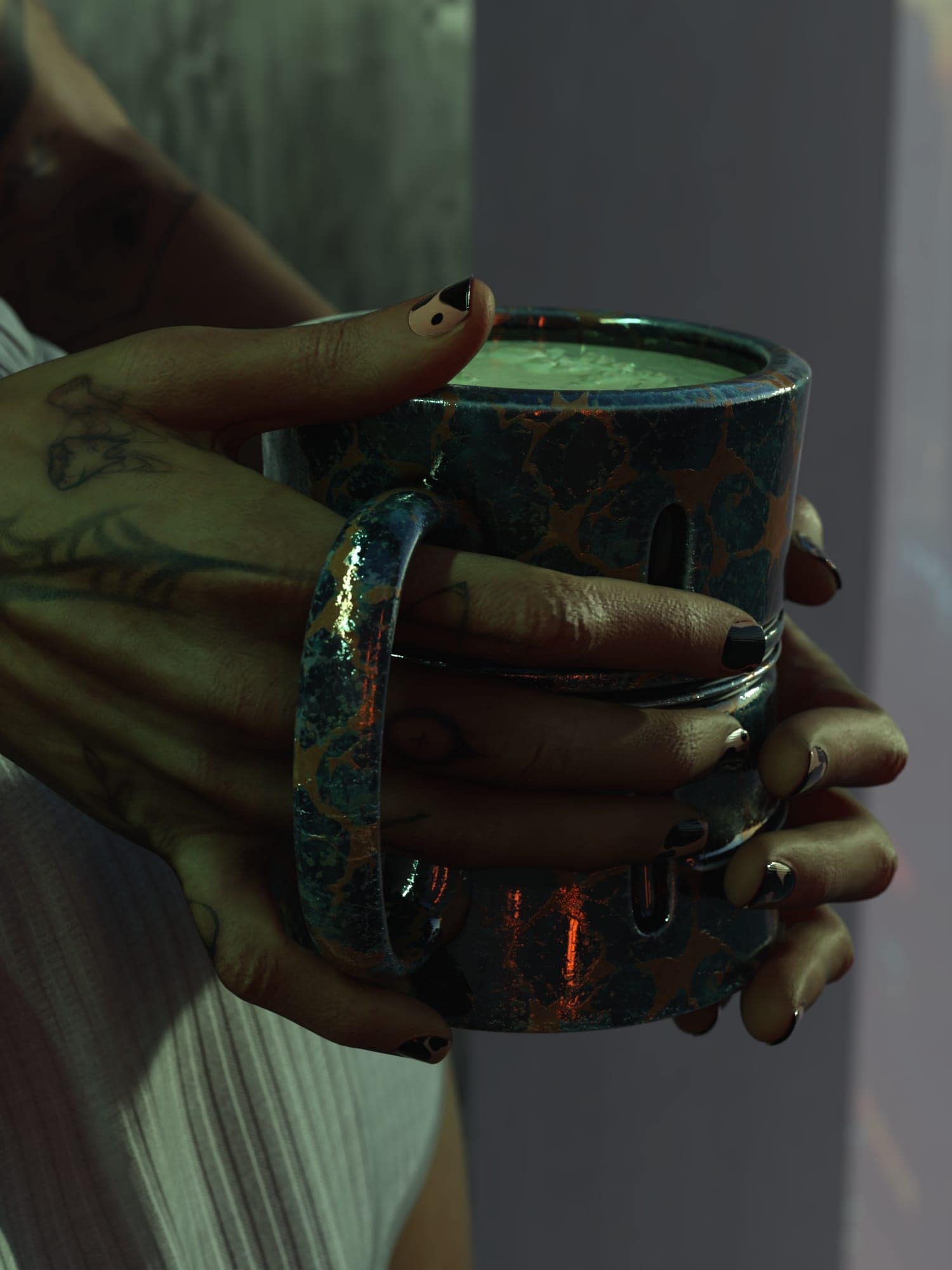 Tattooed hands holding a rustic blue ceramic mug in warm, moody lighting.